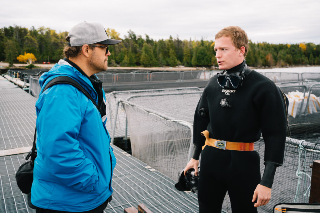 Two men standing and having a conversation. The person on the left is wearing a blue jacket, grey cap and glasses. The person on the right is wearing a black scuba diving suit and has a camera in his hand. Both of them are facing each other and the picture is taken from a side angle.