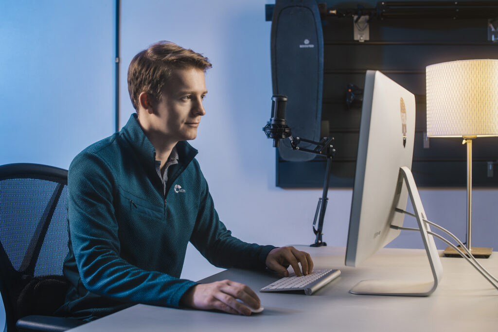 A man in a blue jacket sitting and working on this personal computer. The background is blue and the man is slightly smiling.